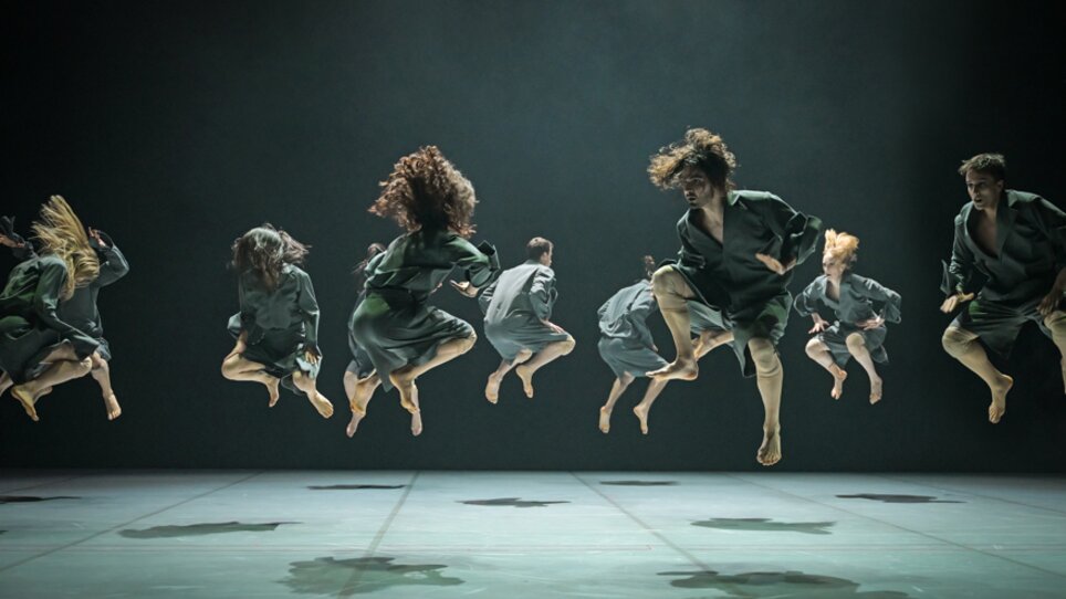 Dancers leap synchronously in the air, energizing and powerful, wearing moss-green attire. | © Andreas J. Etter - Oper Graz