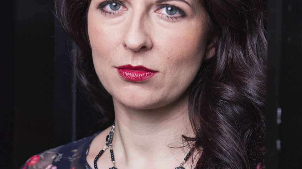 A woman with brown curls and red lipstick, wearing a floral top and a striking necklace. | © Photo-Hagen-Schnauss