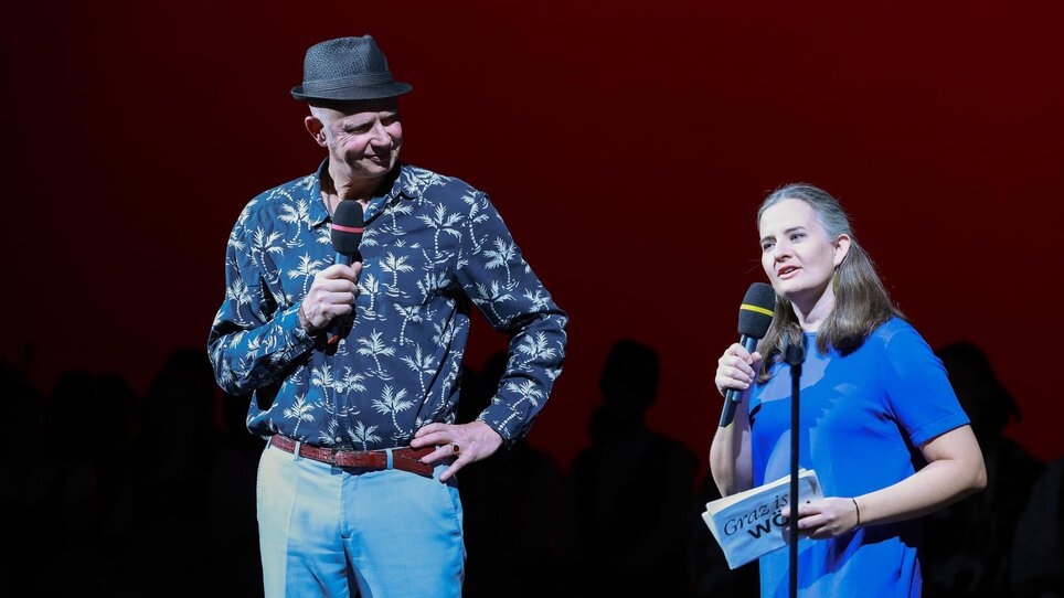 Zwei Moderatoren auf der Bühne, einer mit Mikrofon, der andere spricht. | © Oliver Wolf