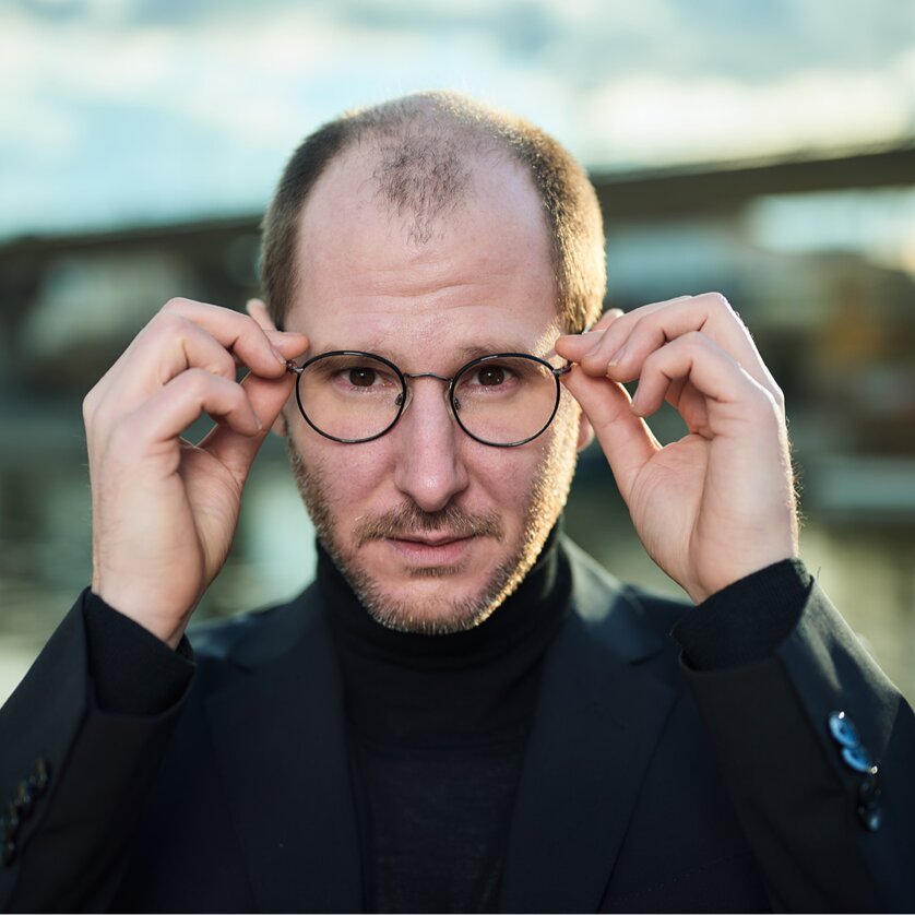 Marco Wanda | © Christopher Mavric Mann mit Brille posiert am Ufer eines Flusses trägt schwarzen Anzug. | © Christopher Mavric