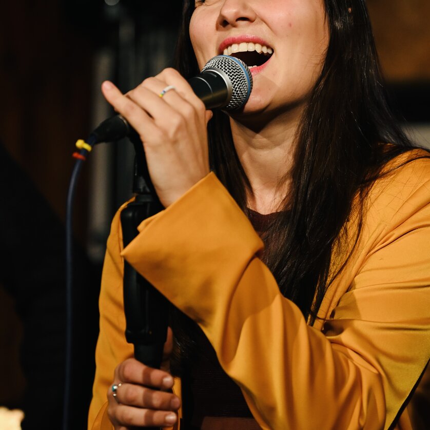 Eine Sängerin mit langen, dunklen Haaren singt ins Mikrofon. | © Bernhard Schaffer