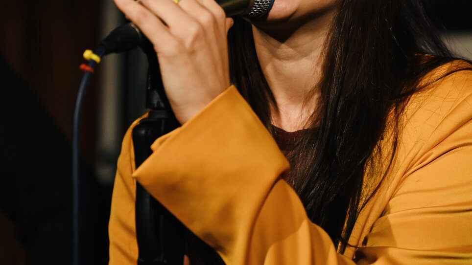 Eine Sängerin mit langen, dunklen Haaren singt ins Mikrofon. | © Bernhard Schaffer