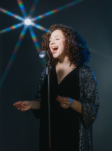 Donna con capelli ricci che canta in un microfono, effetti di luce sullo sfondo. | © Maria Frodl