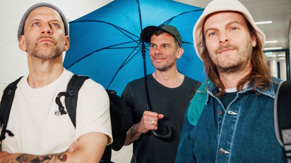 Drei Männer posieren, einer hält einen blauen Regenschirm. | © Paul Ambrusch