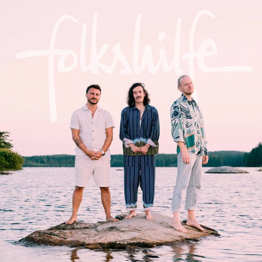 Drei Männer stehen auf einem Felsen im Wasser bei Sonnenuntergang, mit dem Schriftzug 'folkshilfe'. | © Ingo Pertramer