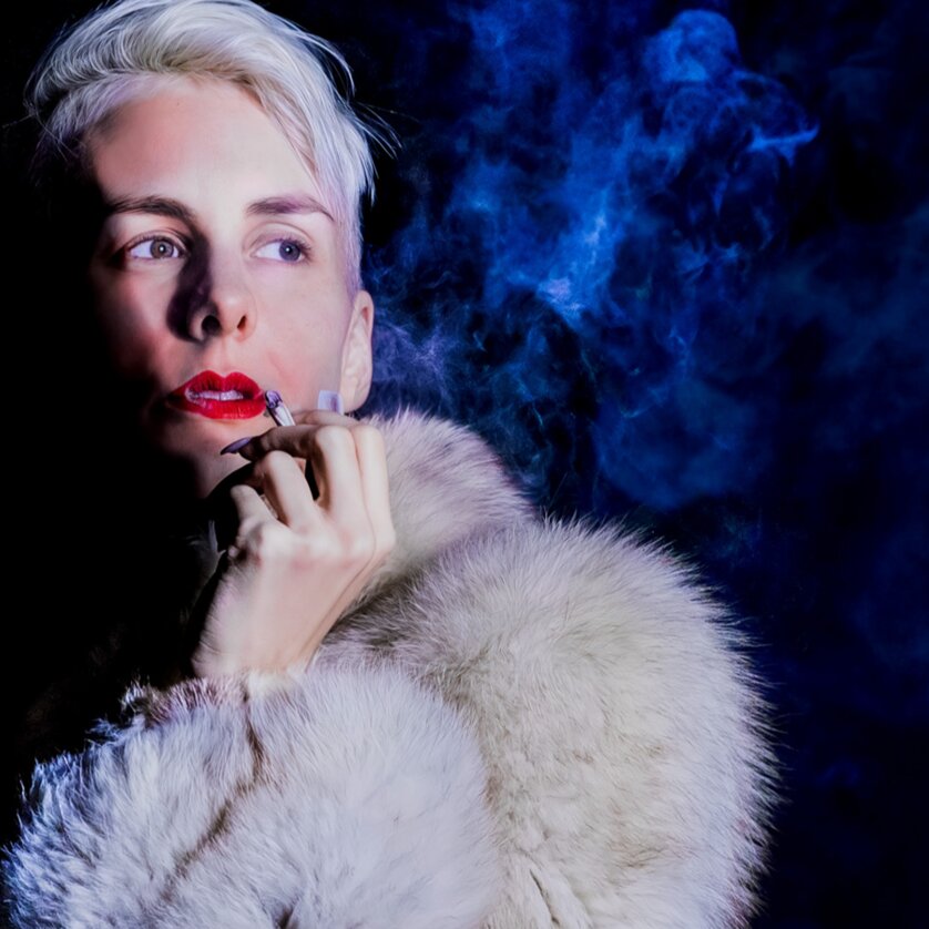 Frau mit blonder Frisur, rotem Lippenstift und Pelzmantel, umgeben von Rauch. | © Jens Doerre