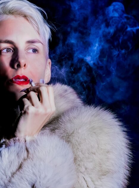 Frau mit blonder Frisur, rotem Lippenstift und Pelzmantel, umgeben von Rauch. | © Jens Doerre