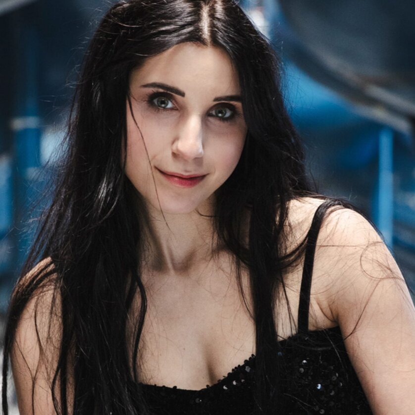 Woman with long dark hair wearing a black top, posing in a modern blue environment. | © Zoe Goldstein