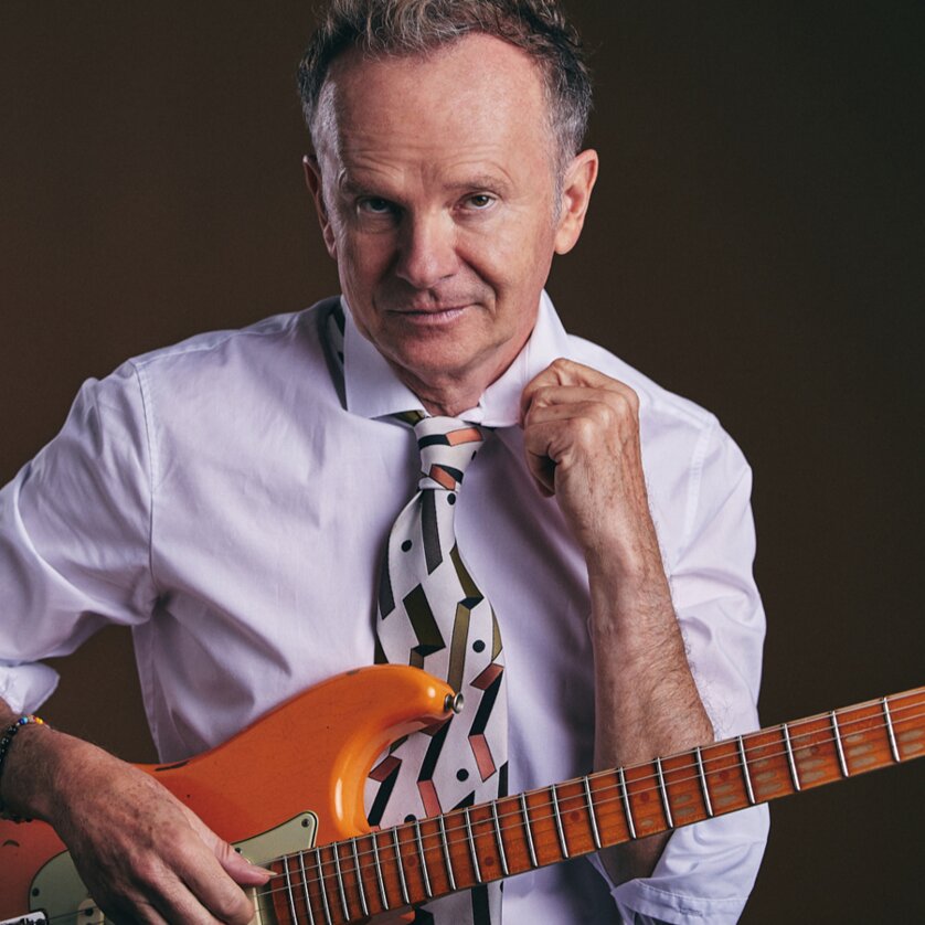 Ein Mann in Weiß mit einer Gitarre und einem gemusterten Hemd und Krawatte. | © Nils Schwarz
