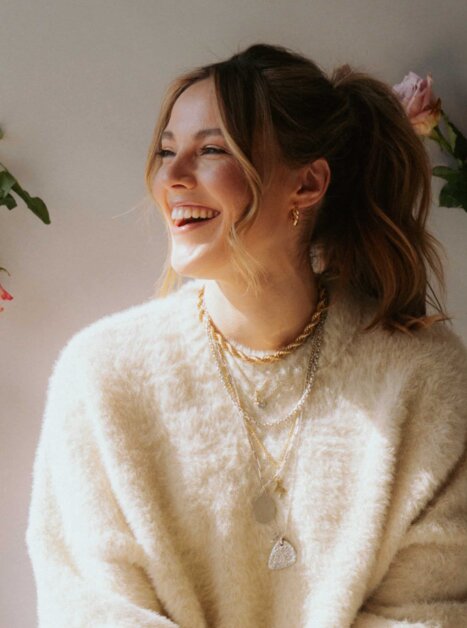 Eine Frau in einem beigen Pullover lächelt vor einer Wand mit Blumen. | © Paul Hüttemann