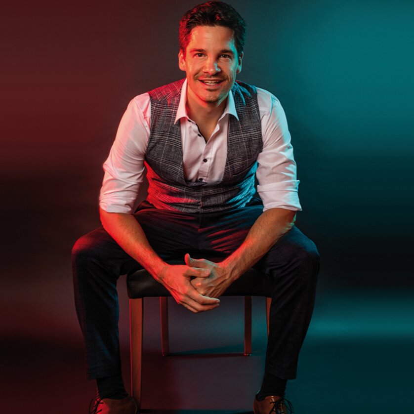 Lächelnder Mann in Weste, sitzt auf Stuhl mit farbigem Hintergrund. | © Mike Wahrlich