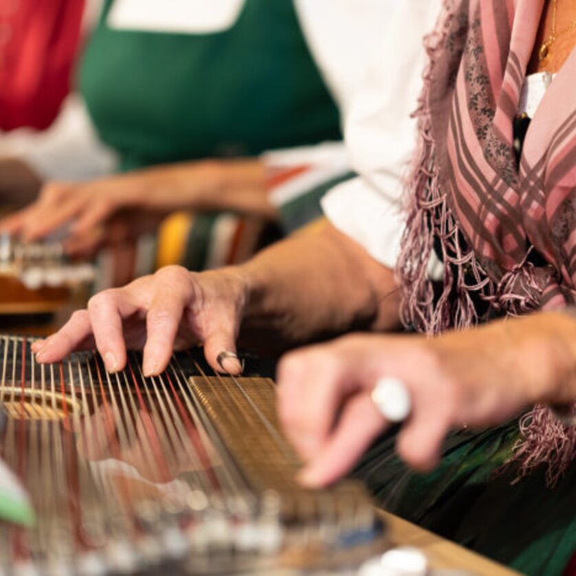 Erster Grazer Zitherverein | © Erster Grazer ZItherverein Hände, die Zither spielen, während Musiker in traditioneller Kleidung sitzen. | © Erster Grazer ZItherverein