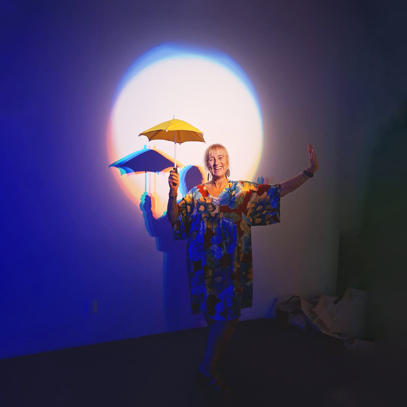 Eine Frau mit Regenschirmen in buntem Lichtspiel steht vor einer Wand. | © Fernanda Meneses