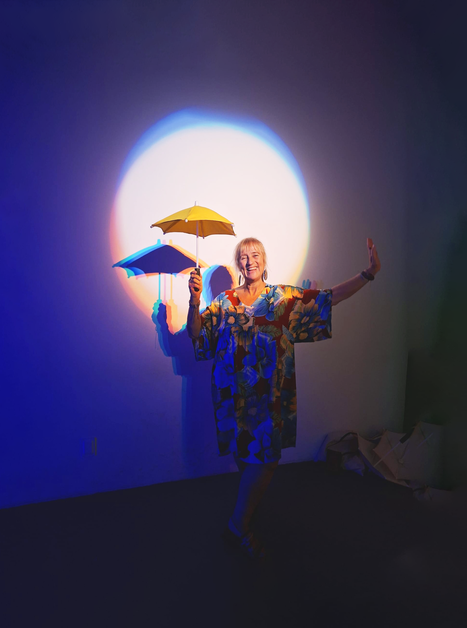 Eine Frau mit Regenschirmen in buntem Lichtspiel steht vor einer Wand. | © Fernanda Meneses