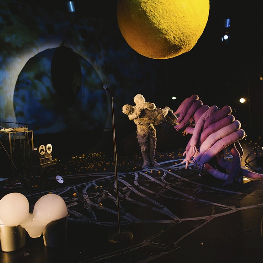 Eine lebendige Inszenierung mit skulpturalen Figuren und bunten Lichtern.