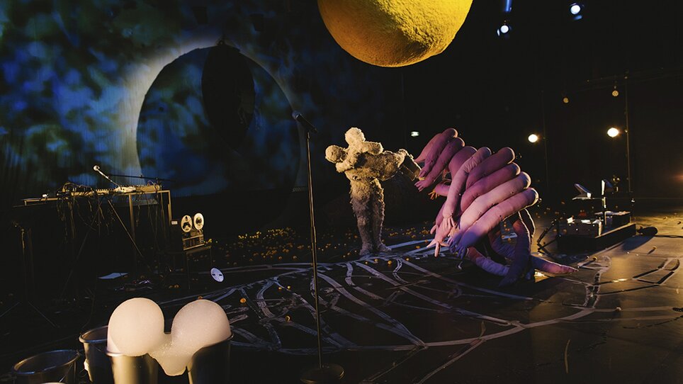 Eine lebendige Inszenierung mit skulpturalen Figuren und bunten Lichtern.