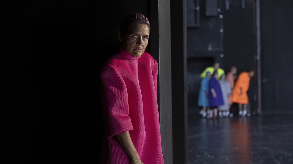 Eine Frau in einem pinken Outfit steht im Schatten, mit einem neutralen Gesichtsausdruck.