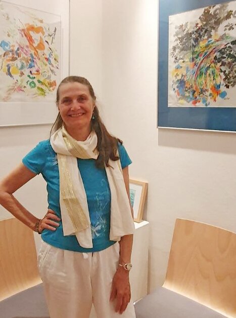 Eine lächelnde Frau steht vor farbigen Kunstwerken an der Wand. | © Sylvia Fruhmann alias LiLa Herderberg
