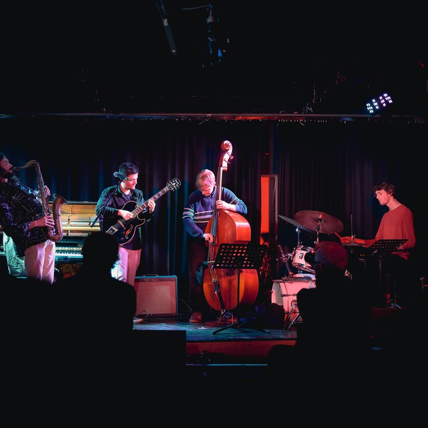 A jazz band plays on a dimly lit stage in front of an audience. | © Kuro Simon