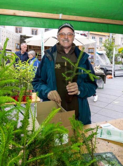 Mann am Waldfest-Stand mit kleinen Tannenbäumchen, umgeben von Besucher:innen. | © Harry Schiffer