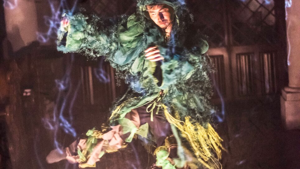 A person in a green costume floats above the ground surrounded by mist. | © Int. Bühnenwerkstatt