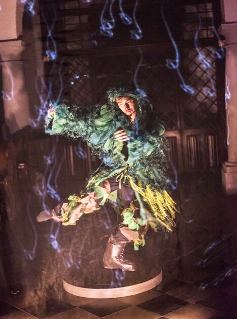 A person in a green costume floats above the ground surrounded by mist. | © Int. Bühnenwerkstatt