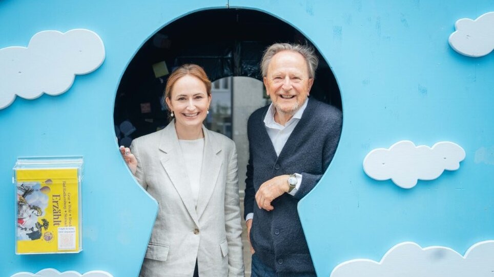 Tessa Erker-Tegetthoff and Folke Tegetthoff are standing in the archway of a blue fairytale castle with white clouds.Two people stand in the archway of a colorful background with clouds. | © Florian Matzi