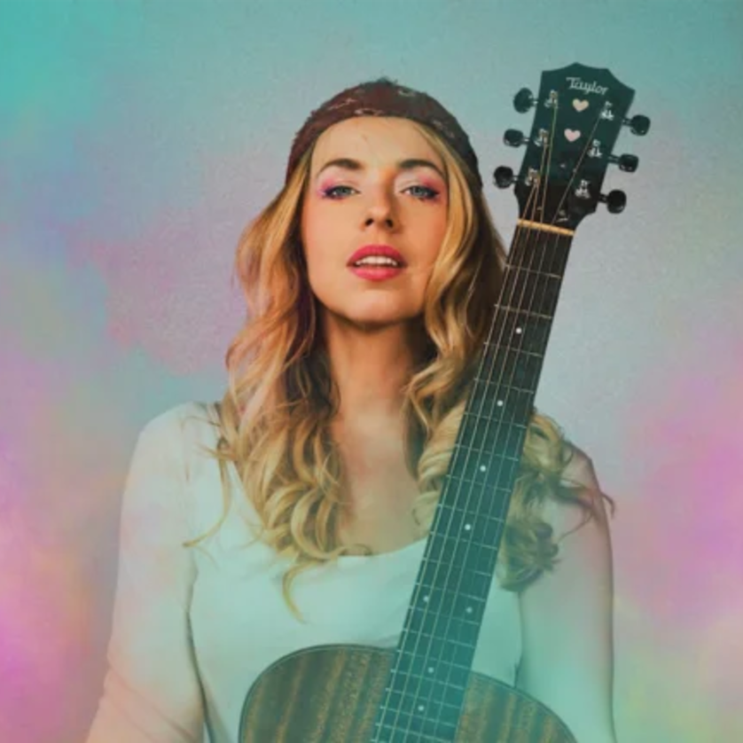 Eine Frau hält eine Gitarre, stehend vor einem bunten Hintergrund. | © Christian Verch