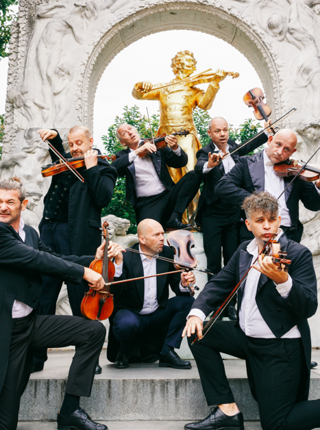 Eine Gruppe von Musikern posiert mit Geigen vor einer Statue. | © Maria Frodl