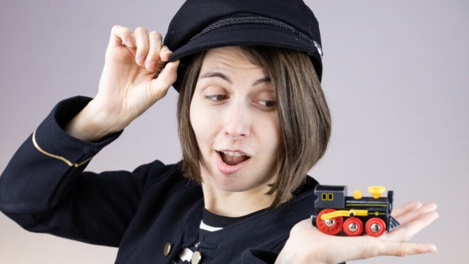 Eine Person in Uniform zeigt erstaunt auf eine kleine Spielzeuglok. | © Magda Leeb