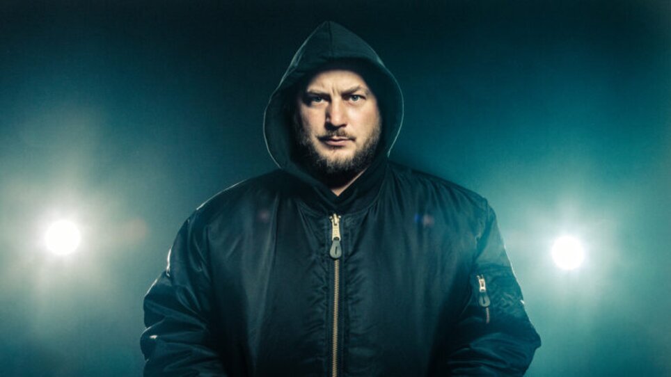 Ein Mann in einer dunklen Jacke mit Kapuze steht leidenschaftlich vor einem beleuchteten Hintergrund. | © Antonia Mayer