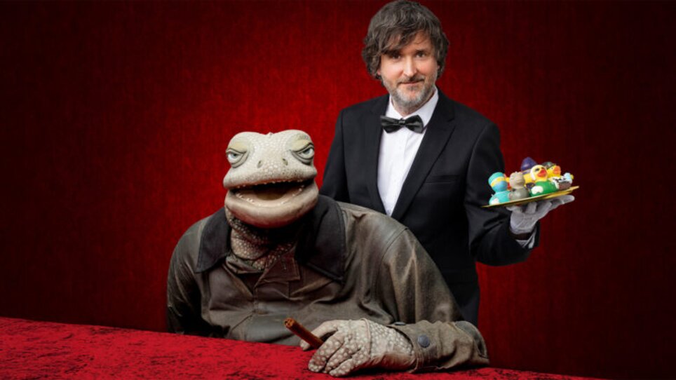 Ein Frosch im Anzug sitzt an einem Tisch, während ein Kellner einen Teller serviert. | © Paul Jackel