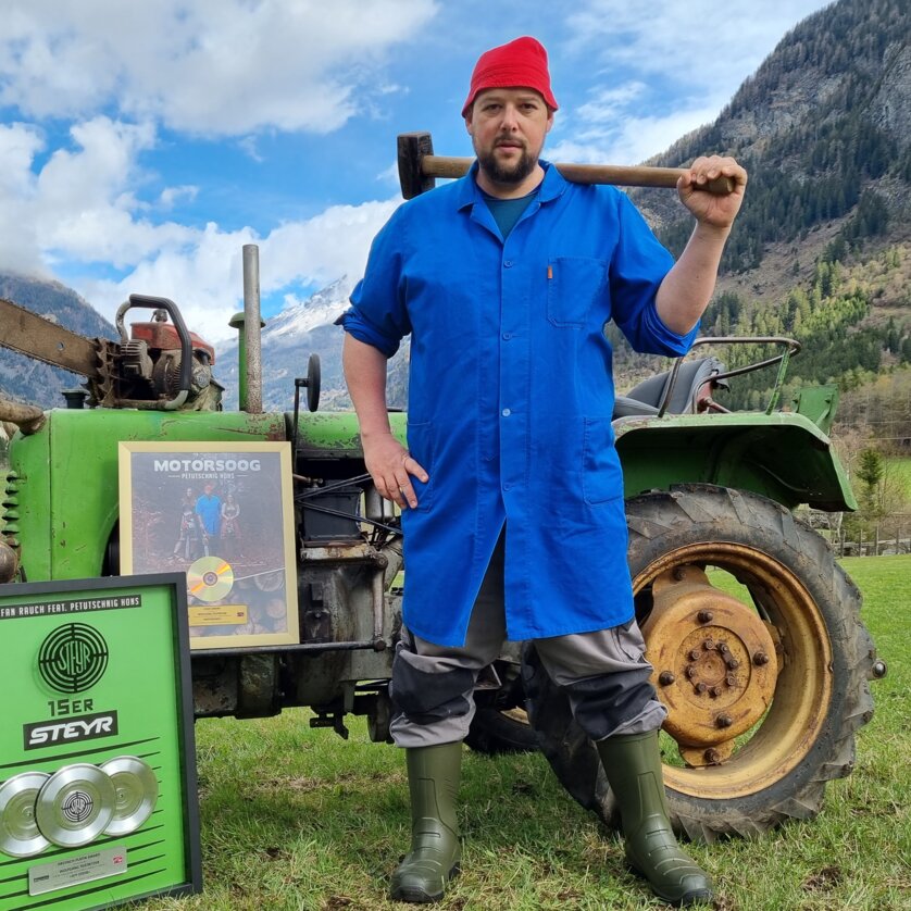 Ein Mann in blauer Jacke steht mit einer Axt neben einem Traktor auf einer Wiese. | © Petutschnig Hons
