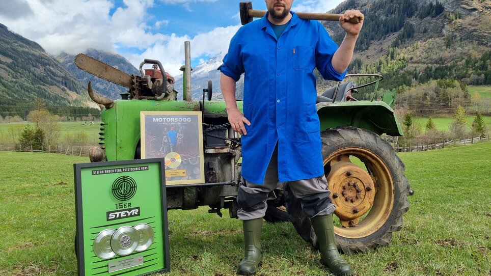 Ein Mann in blauer Jacke steht mit einer Axt neben einem Traktor auf einer Wiese. | © Petutschnig Hons