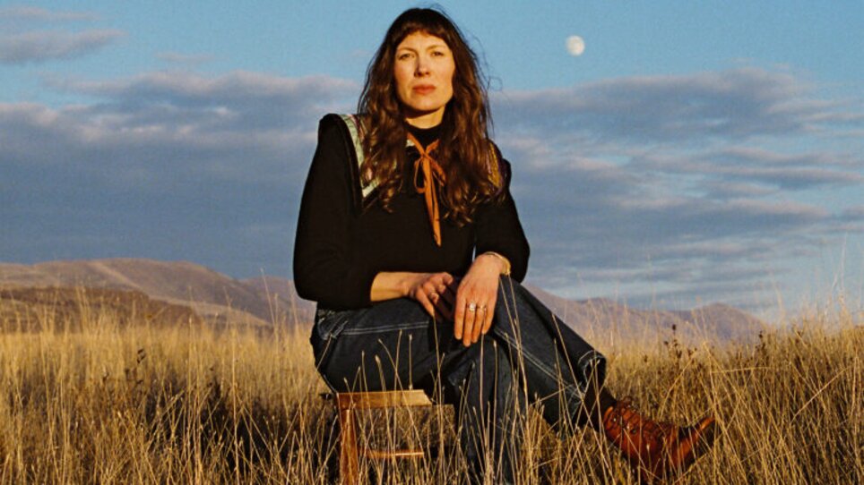 Frau sitzt auf einem Hocker in einem Feld, der Mond ist im Hintergrund sichtbar. | © Kelsey Gallagher