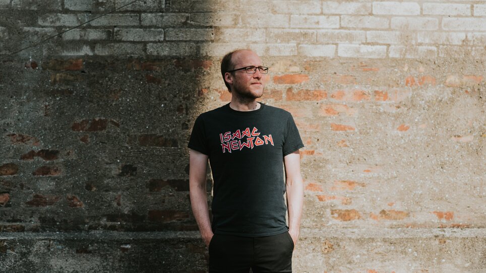 Ein Mann steht neben einer Backsteinmauer in einem T-Shirt mit Isaac Newton Aufdruck. | © Franzi Schädel - CC-BY-SA 4.0