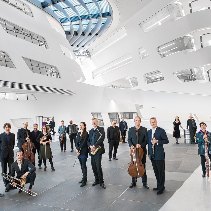 a group of musicians are in a building with modern interior with curved wall elements and glass panels | © Tina Herzl