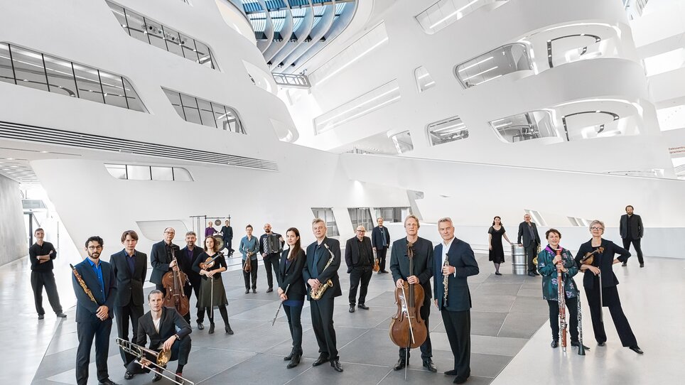 a group of musicians are in a building with modern interior with curved wall elements and glass panels | © Tina Herzl