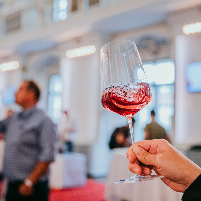 Wine glass filled with rosé | © Johanna Lamprecht A wine glass with shimmering red wine is held up to the camera. The wine swirls in the glass, people can be recognised blurred in the background. The atmosphere is festive and stylish. | © Johanna Lamprecht