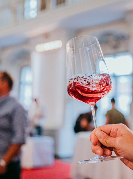 A wine glass with shimmering red wine is held up to the camera. The wine swirls in the glass, people can be recognised blurred in the background. The atmosphere is festive and stylish. | © Johanna Lamprecht
