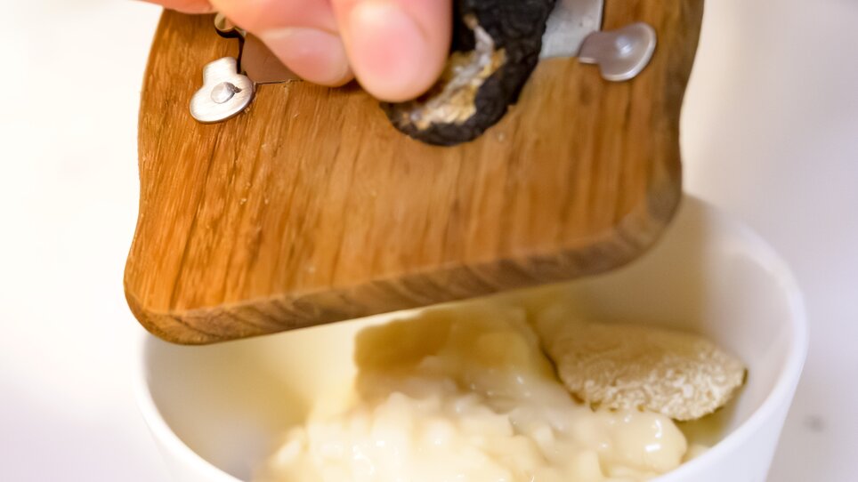 Eine Hand reibt eine schwarze Trüffel über cremigem Risotto in einer weißen Schüssel. | © ivents Kulturagentur
