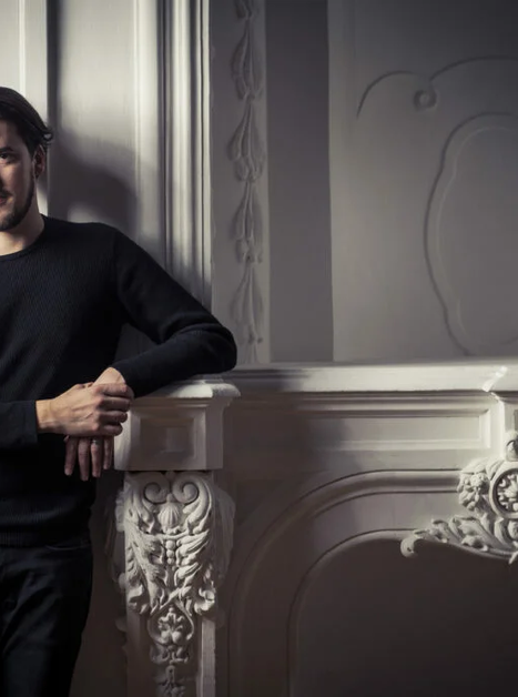 Junger Mann in schwarzem Pullover steht neben einer dekorativen Wand. | © Guido Werner