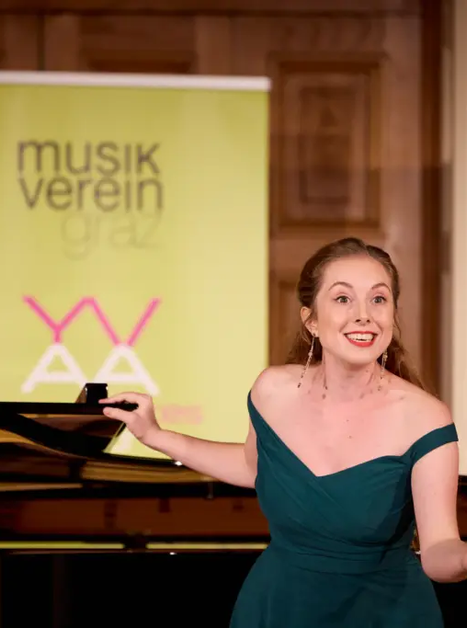 A singer in a green dress stands in front of a piano, smiling. | © Kurt Remling