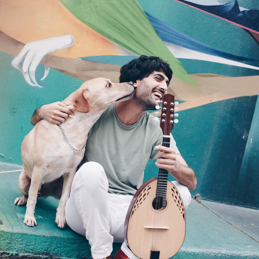 Ein Mann sitzt mit Hund und Mandoline vor einem bunten Wandgemälde. | © Juliana Farinha