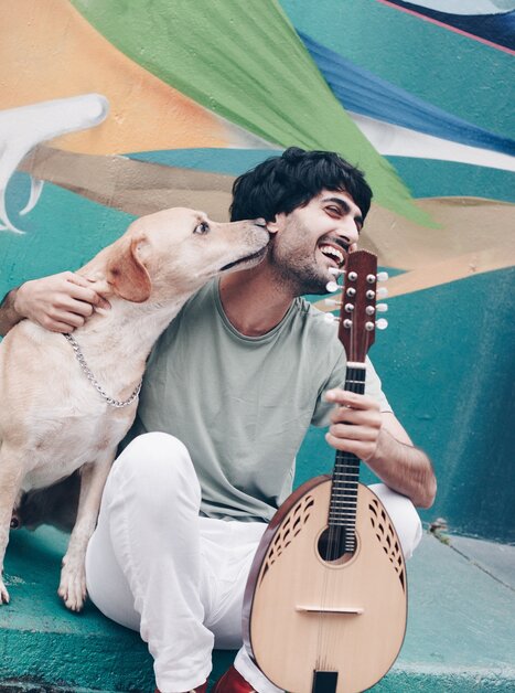 Ein Mann sitzt mit Hund und Mandoline vor einem bunten Wandgemälde. | © Juliana Farinha
