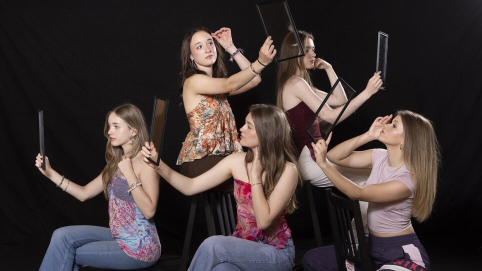Fünf junge Frauen sitzen mit Handspiegeln und blicken hinein vor schwarzem Hintergrund. | © Peter Manninger