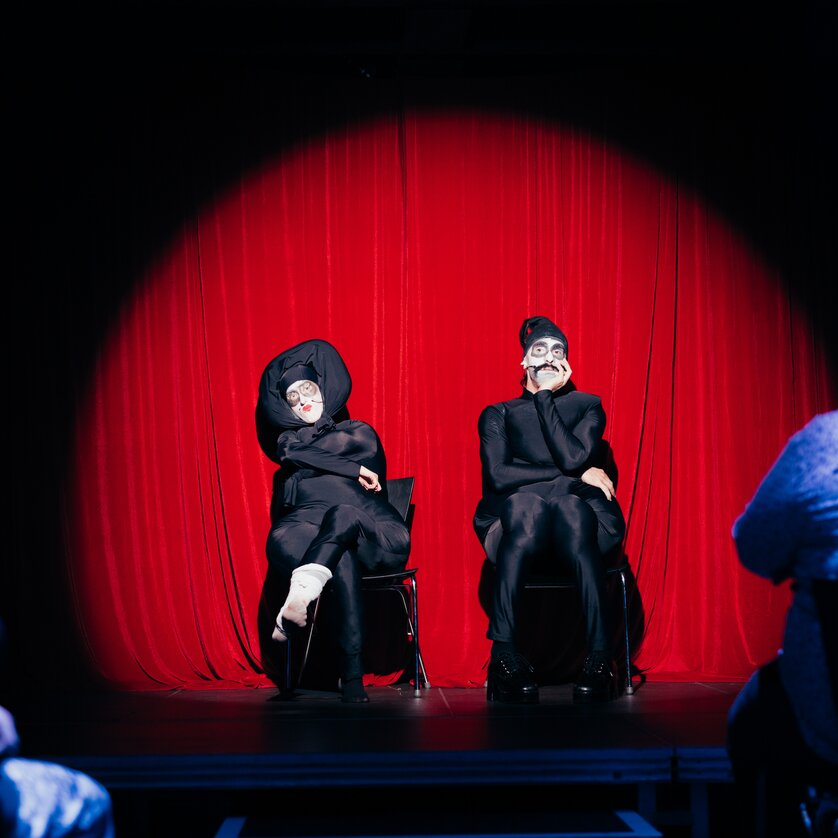 Zwei Performer in Masken auf der Bühne vor einem roten Vorhang. | © Daniel Kindler