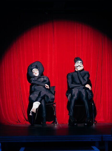 Two performers in masks on stage in front of a red curtain. | © Daniel Kindler