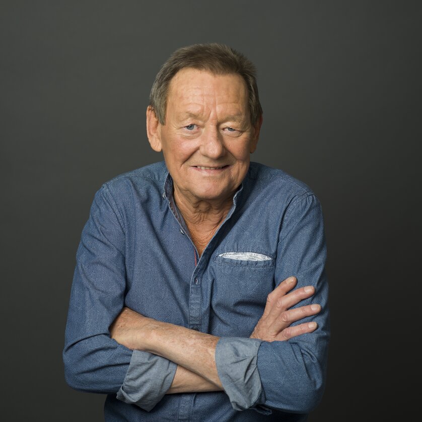 Wolfgang Ambros, österreichischer Musiker und Austropop-Legende, trägt ein blaues Jeans-Hemd, steht mit verschränkten Armen und lächelt in die Kamera.