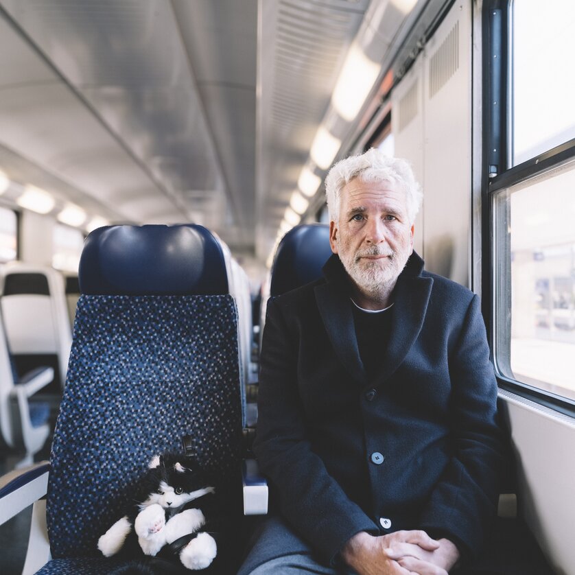 Ein älterer Mann sitzt neben einer schwarzen und weißen Katze im Zug. | © Ingo Pertramer